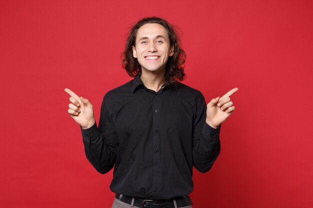 Knappe stijlvolle jonge krullend langharige man in zwart shirt poseren geïsoleerd op rode muur achtergrond studio portret. Mensen oprechte emoties levensstijl concept Mock up wijzende vinger hand op kopie ruimte