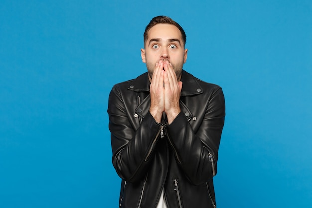 Knappe stijlvolle jonge ongeschoren man in zwart lederen jas wit t-shirt op zoek camera geïsoleerd op blauwe muur achtergrond studio portret. mensen oprechte emoties levensstijl concept. bespotten kopie ruimte