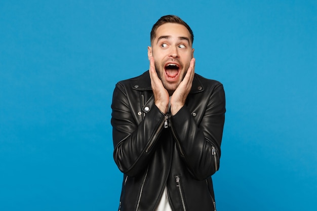 Knappe stijlvolle jonge ongeschoren man in zwart lederen jas wit t-shirt op zoek camera geïsoleerd op blauwe muur achtergrond studio portret. Mensen oprechte emoties levensstijl concept. Bespotten kopie ruimte