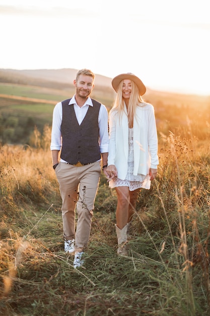 Knappe stijlvolle man in shirt, vest en broek en mooie boho vrouw in jurk, jas en hoed wandelen in het veld