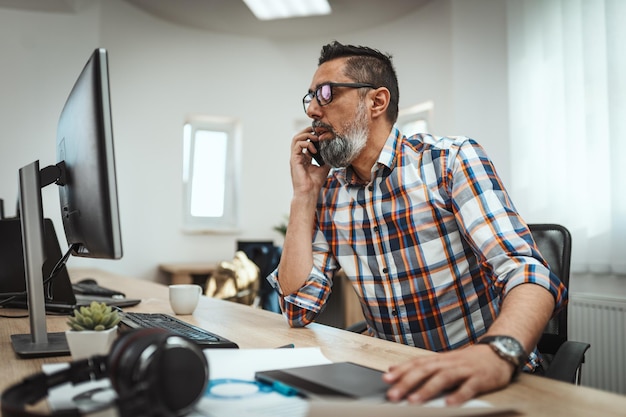 Knappe succesvolle ondernemer praat op een mobiele telefoon terwijl hij op kantoor werkt.