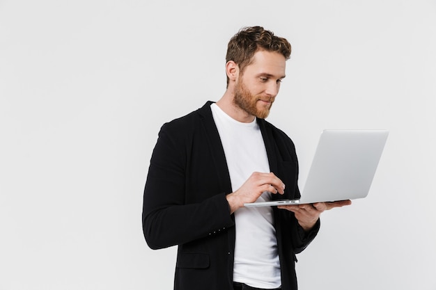 knappe tevreden man in jas glimlachend en met behulp van laptop geïsoleerd over witte muur