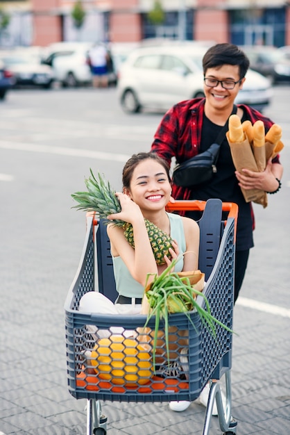 Knappe Vietnamese man houdt papieren zakken met voedsel duwen voor hem winkelwagentje met zijn gelukkige mooie vriendin erin. Grappige familie winkelen.