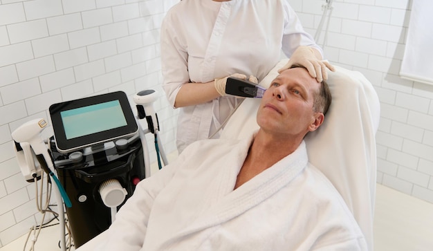 Knappe volwassen Europese man in witte badstof badjas die mesotherapie voor het gezicht krijgt in een modern wellness-spacentrum. Hardware moderne cosmetologie, mesotherapie, verjonging en anti-aging concept