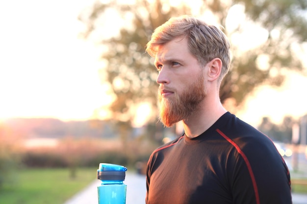 Knappe volwassen man die water uit een fitnessfles drinkt terwijl hij buiten staat, bij zonsondergang of zonsopgang. loper