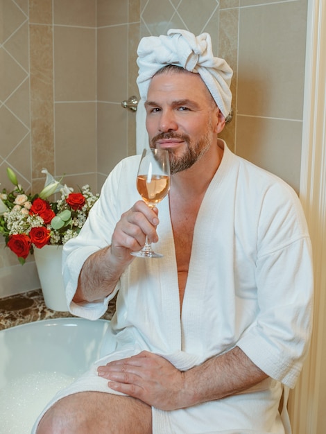 knappe volwassen man homo in witte badjas en handdoek op zijn hoofd zittend op badkamer met schuim