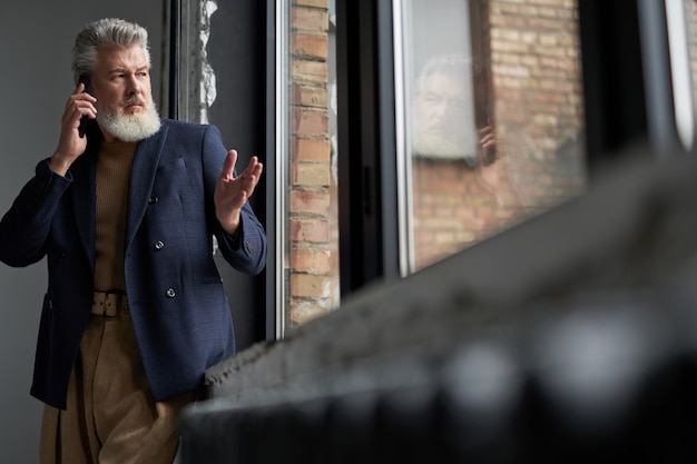 Knappe volwassen man in zakelijke vrijetijdskleding die uit een groot raam kijkt, telefoneert met een smartphone terwijl hij in het interieur van de loft staat. Levensstijl, mensenconcept