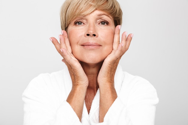 knappe volwassen vrouw met kort blond haar die een witte huisjas draagt en haar gezicht aanraakt dat over een witte muur wordt geïsoleerd