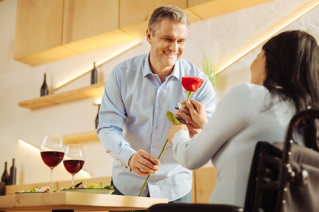 Knappe, vrolijke, goed gebouwde man die lacht en een rode bloem geeft aan zijn geliefde donkerharige gehandicapte vrouw tijdens een romantisch diner