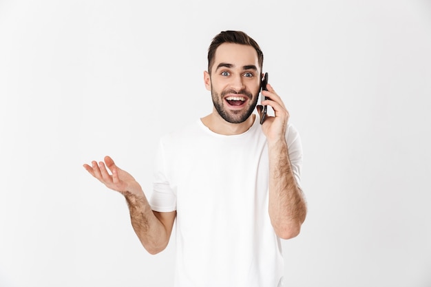 Knappe vrolijke man met een leeg t-shirt dat geïsoleerd staat over een witte muur, met behulp van mobiele telefoon