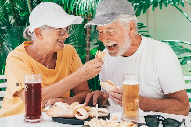 Knappe, vrolijke oudere echtpaar in hoeden zitten aan een kroegtafel en eten broodjes en genieten van koude bieren.