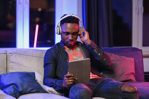 Knappe vrolijke stijlvolle Afro-Amerikaanse met koptelefoon op zijn nek ontspannen op de zachte bank thuis en browsen apps op tablet pc met een gelukkige glimlach