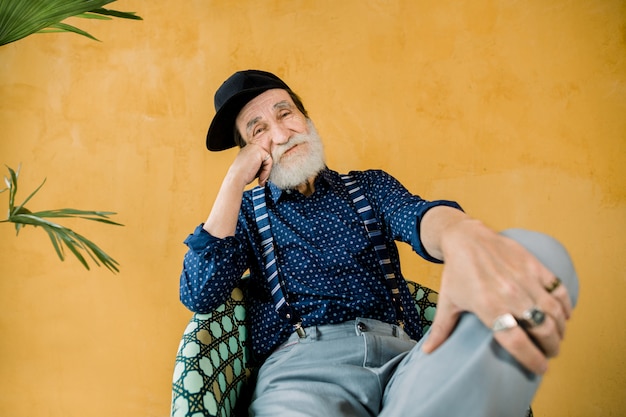 Knappe vrolijke trendy senior man met goed verzorgde baard, gekleed in donkerblauw shirt, bretels, grijze broek en zwarte hipster cap, zittend in stoel in studio voor gele muur