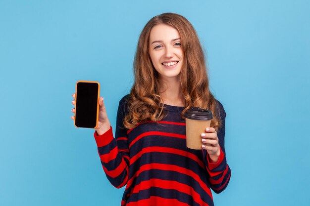 Knappe vrouw met golvend haar die een gestreepte casual stijltrui draagt die koffie houdt om te gaan en een smartphone met leeg zwart scherm. Indoor studio opname geïsoleerd op blauwe achtergrond.