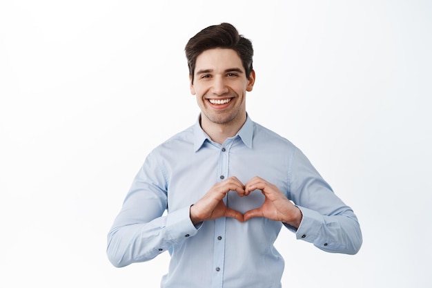 Knappe zakenman die een hartteken toont en glimlacht, ik hou van je als een gebaar, staande op een witte achtergrond in een kantoorshirt