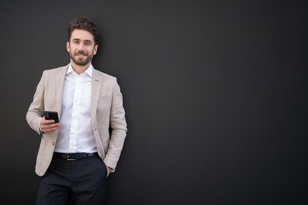 Knappe zakenman die naast een zwarte muur staat met een smartphone in de hand