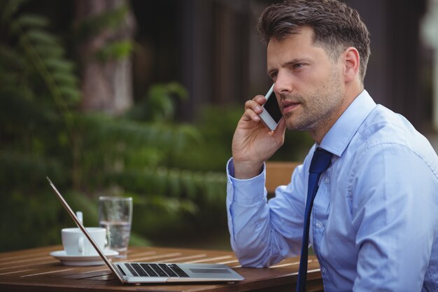 Knappe zakenman die op mobiele telefoon spreekt terwijl het gebruiken van laptop