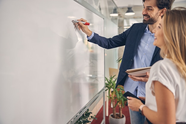 Knappe zakenman die op whiteboard schrijft en glimlacht terwijl zijn vrouwelijke collega smartphone vasthoudt