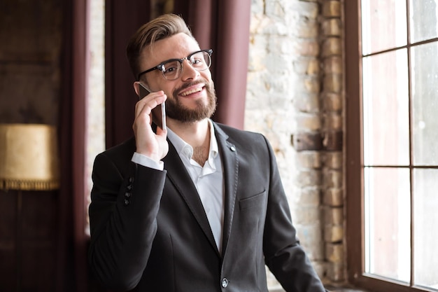 Knappe zakenman in pak en bril die aan de telefoon spreekt in kantoor bij het raam