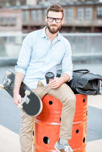 Knappe zakenman met een koffiepauze zittend met skateboard op het dak op de industriële achtergrond