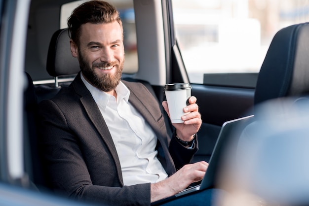 Knappe zakenman zit met koffie om op de achterbank van de auto te gaan