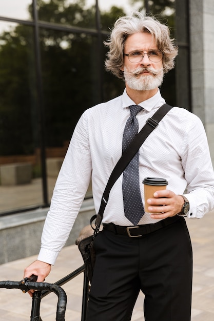 Knappe zelfverzekerde bebaarde zakenman die een pak draagt, een aktetas draagt die buiten staat met een fiets, met afhaalkoffie