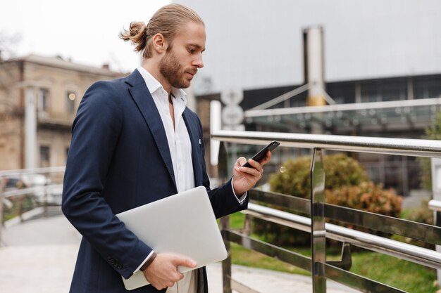 Knappe zelfverzekerde jonge, bebaarde zakenman die buiten op straat staat en mobiele telefoon gebruikt terwijl hij een laptop draagt