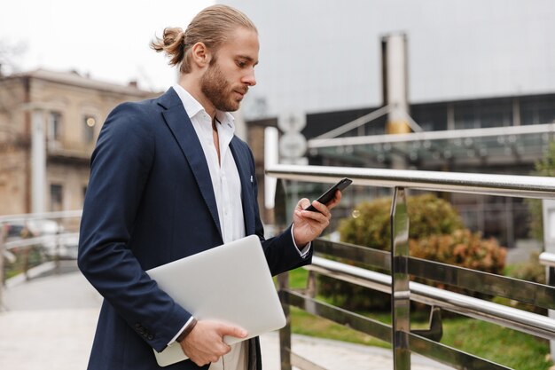 Foto knappe zelfverzekerde jonge bebaarde zakenman die buiten op straat staat en mobiele telefoon gebruikt terwijl hij een laptop draagt