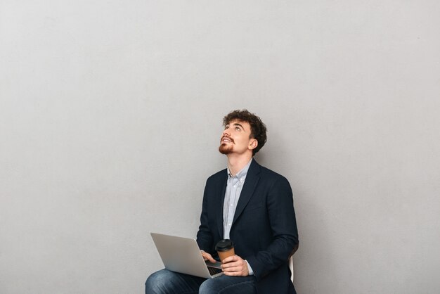 Knappe zelfverzekerde jonge zakenman die een jas draagt die in een stoel zit die over grijs wordt geïsoleerd, die aan laptop computer werkt, afhaalmaaltijdenkoffie houdt