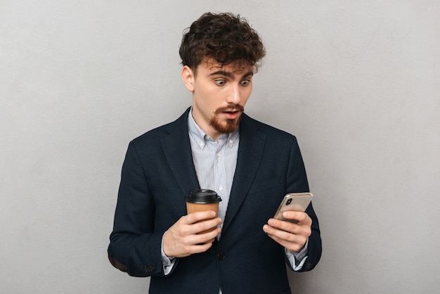 Knappe zelfverzekerde jonge zakenman die een jas draagt die zich geïsoleerd over grijs bevindt, die mobiele telefoon gebruikt, die kop afhaalkoffie houdt