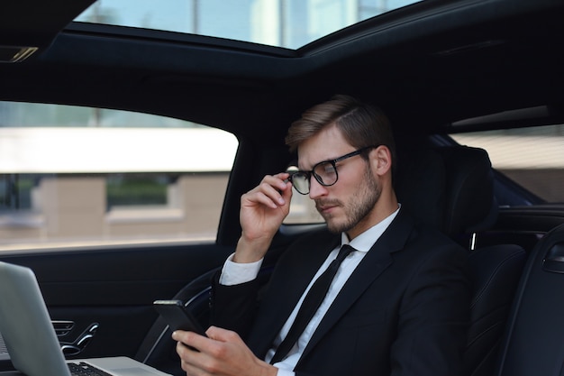 Knappe zelfverzekerde man in volledig pak die naar zijn smartphone kijkt terwijl hij in de auto zit en een laptop gebruikt.