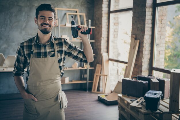 knappe zelfverzekerde man meester bedrijf elektrische draadloze boor vriendelijk lachend klaar om te beginnen reparatie eigen houten bedrijf industrie studio houtwerk winkel garage binnenshuis