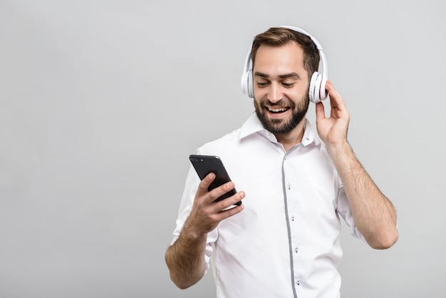 Knappe zelfverzekerde zakenman die een pak draagt dat geïsoleerd staat over een grijze muur, luistert naar muziek met oortelefoons en mobiele telefoon
