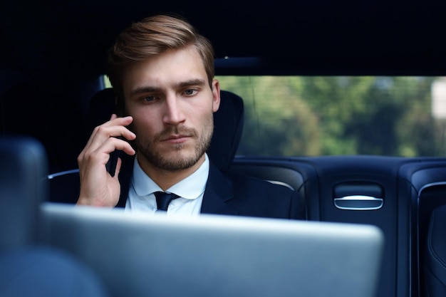 Knappe zelfverzekerde zakenman in pak die op een smartphone praat en met een laptop werkt terwijl hij in de auto zit