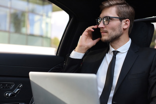 Knappe zelfverzekerde zakenman in pak praten op slimme telefoon en werken met behulp van laptop zittend in de auto.