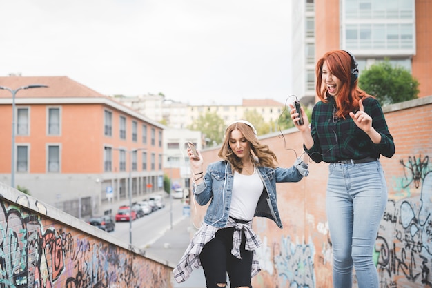 Knee figure of two young handsome caucasian blonde and redhead straight hair women dancing in the city listening music with headphones and smartphone, laughing - music, fun, freedom concept