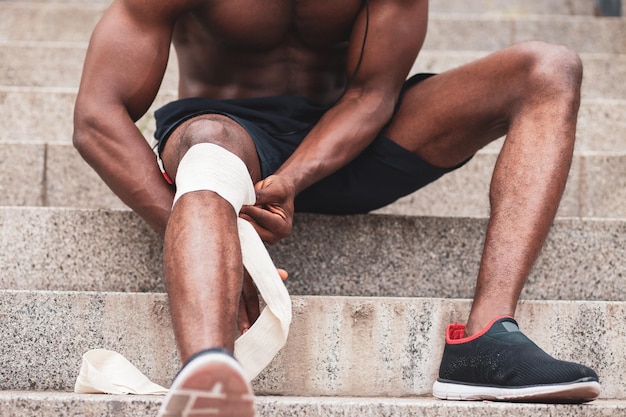 Knee injury concept afro american guy bandaging his leg with a\
bandage