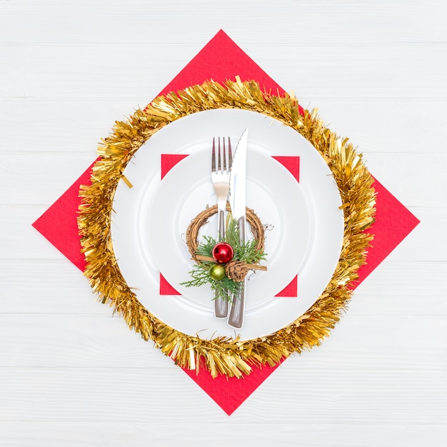 Нож и вилка в белой тарелке на красной салфетке украшены рождественским венком на белом столе