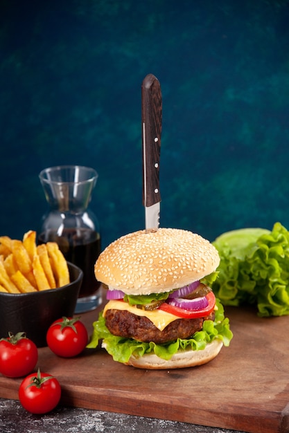 Knife in tasty meat sandwich sauce ketchup fries on wooden board and tomatoes with stem on dark blue surface