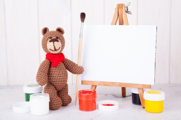 Knitted brown bear with brush and paint near the easel. Handwork, creativity. Amigurumi. Postcard