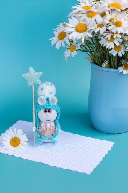 Knitted bunny in a hat on a blue background bouquet of daisies in the background