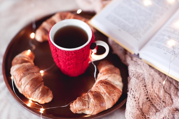 Knitted cup of black tea with homemade croissants and open book staying on wooden tray in bed closeup Winter season Good morning