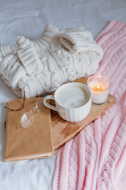 Maglione lavorato a maglia, plaid rosa, candela, tazza da cappuccino, libro, bicchieri sul letto. accogliente colazione. inverno. autunno.