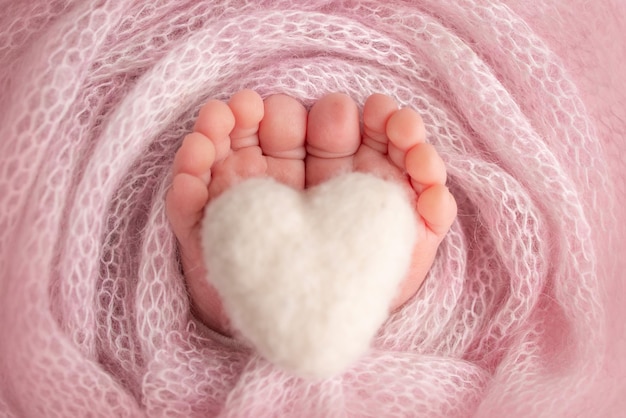 Knitted white heart in the legs of a baby Soft feet of a new born in a pink wool blanket Closeup of toes heels and feet of a newborn Macro photography the tiny foot of a newborn baby