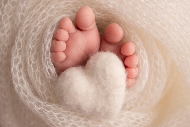 Knitted white heart in the legs of a baby Soft feet of a new born in a white wool blanket Closeup of toes heels and feet of a newborn Macro photography the tiny foot of a newborn baby