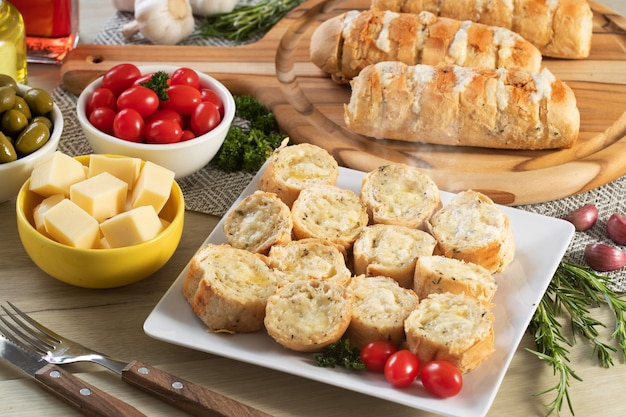 Knoflookbrood in witte vierkante plaat op tafel met kaas, rozemarijn, olijven en kerstomaatjes.