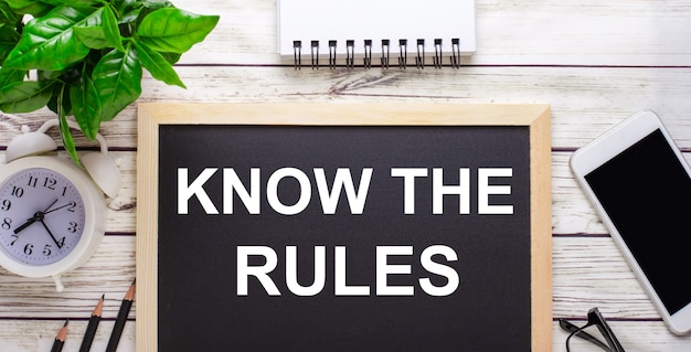 KNOW THE RULES written on a black surface near pencils, a smartphone, a white notepad and a green plant in a pot