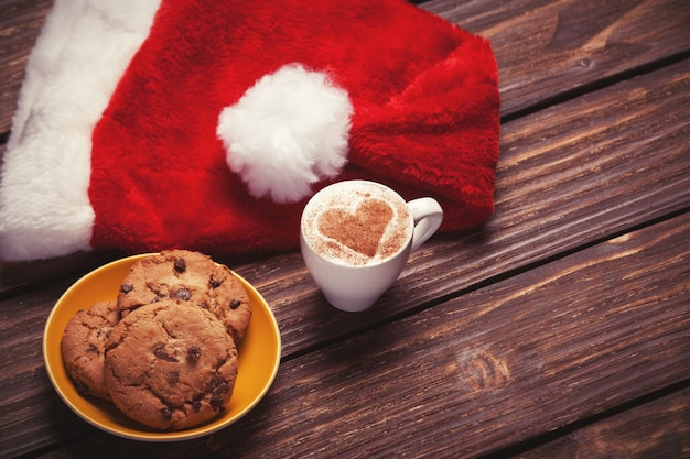 Koekje en kopje koffie met kerstmuts op houten tafel.