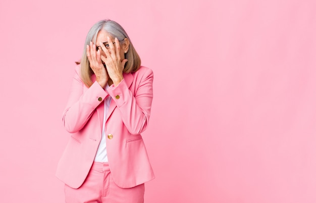 Koele vrouw van middelbare leeftijd die zich bang of beschaamd voelt, gluurt of spioneert met ogen half bedekt met handen over de muur