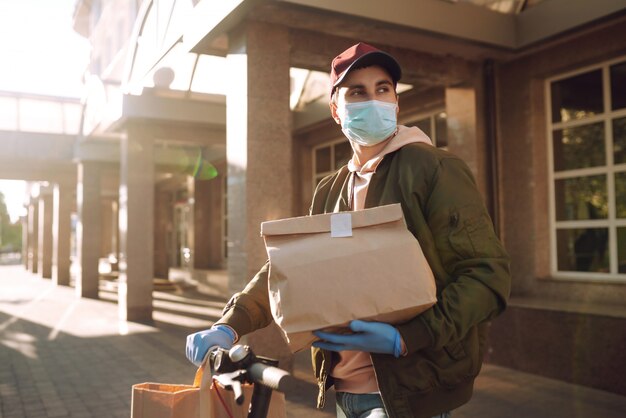 Koerier in beschermend masker en medische handschoenen op een scooter levert ambachtelijke papieren pakket met voedsel. Bezorgservice onder quarantaine, ziekte-uitbraak, coronavirus covid-19 pandemische aandoeningen.